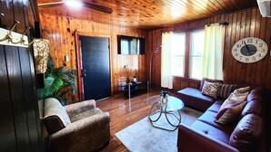 Een gezellige woonkamer met houten wanden en plafond, een bank, een fauteuil, een bijzettafeltje en een klok aan de muur.