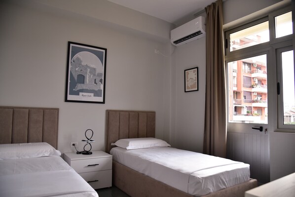 Ein Hotelzimmer mit zwei Betten, einem Nachttisch, einem gerahmten Bild eines Gebäudes und einem Fenster mit Blick auf andere Gebäude.