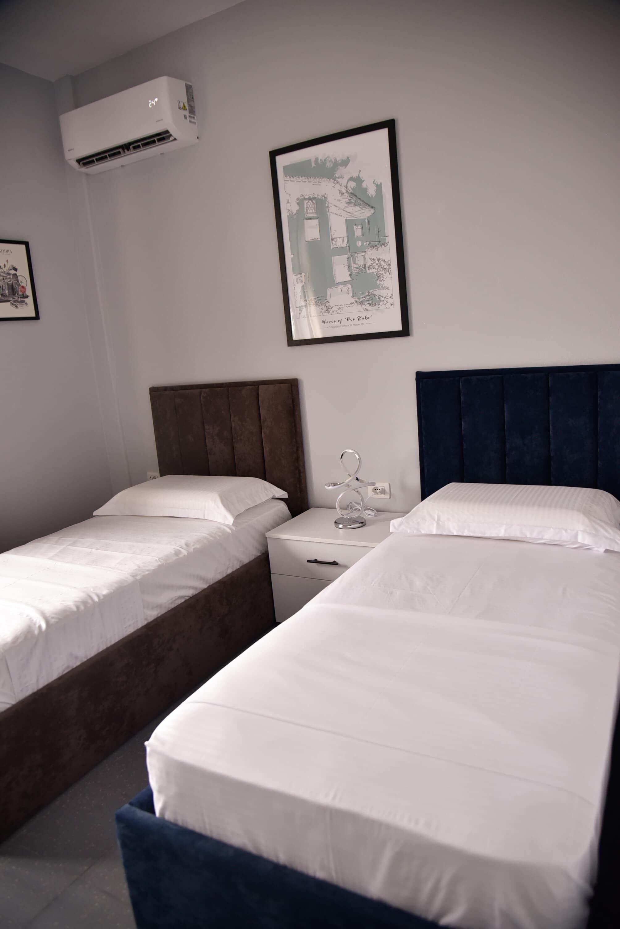 A hotel room with two beds, a nightstand, a framed picture, and an air conditioning unit.