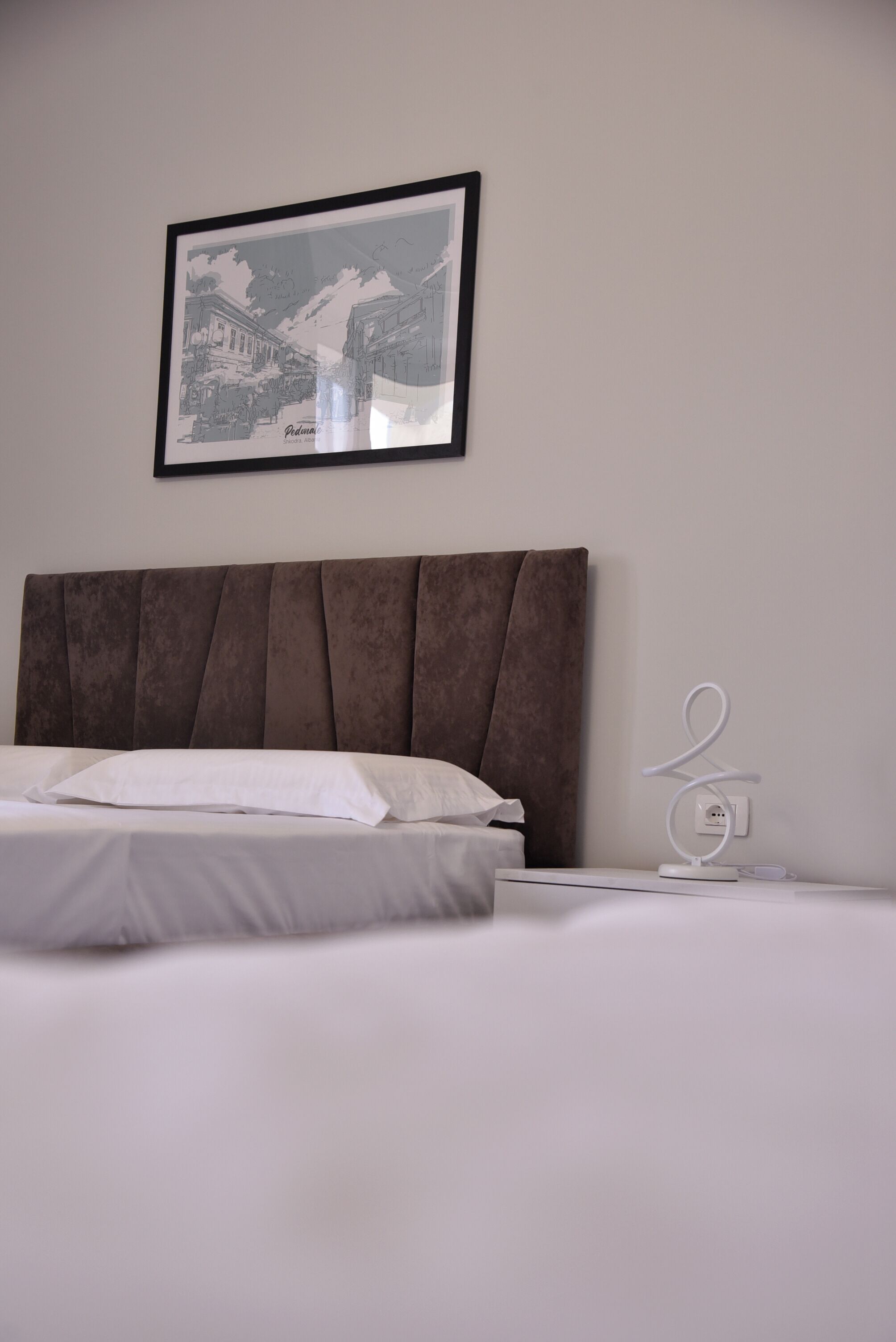 Un lit bien fait, avec du linge de lit blanc, une tête de lit marron recouverte de tissu et un tableau encadré accroché au mur.