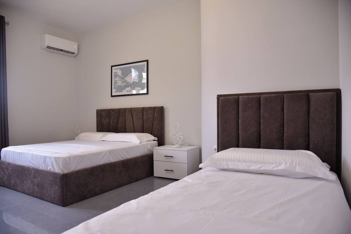 A modern hotel room with two beds, a bedside table, a wall-mounted air conditioner, and a framed picture.