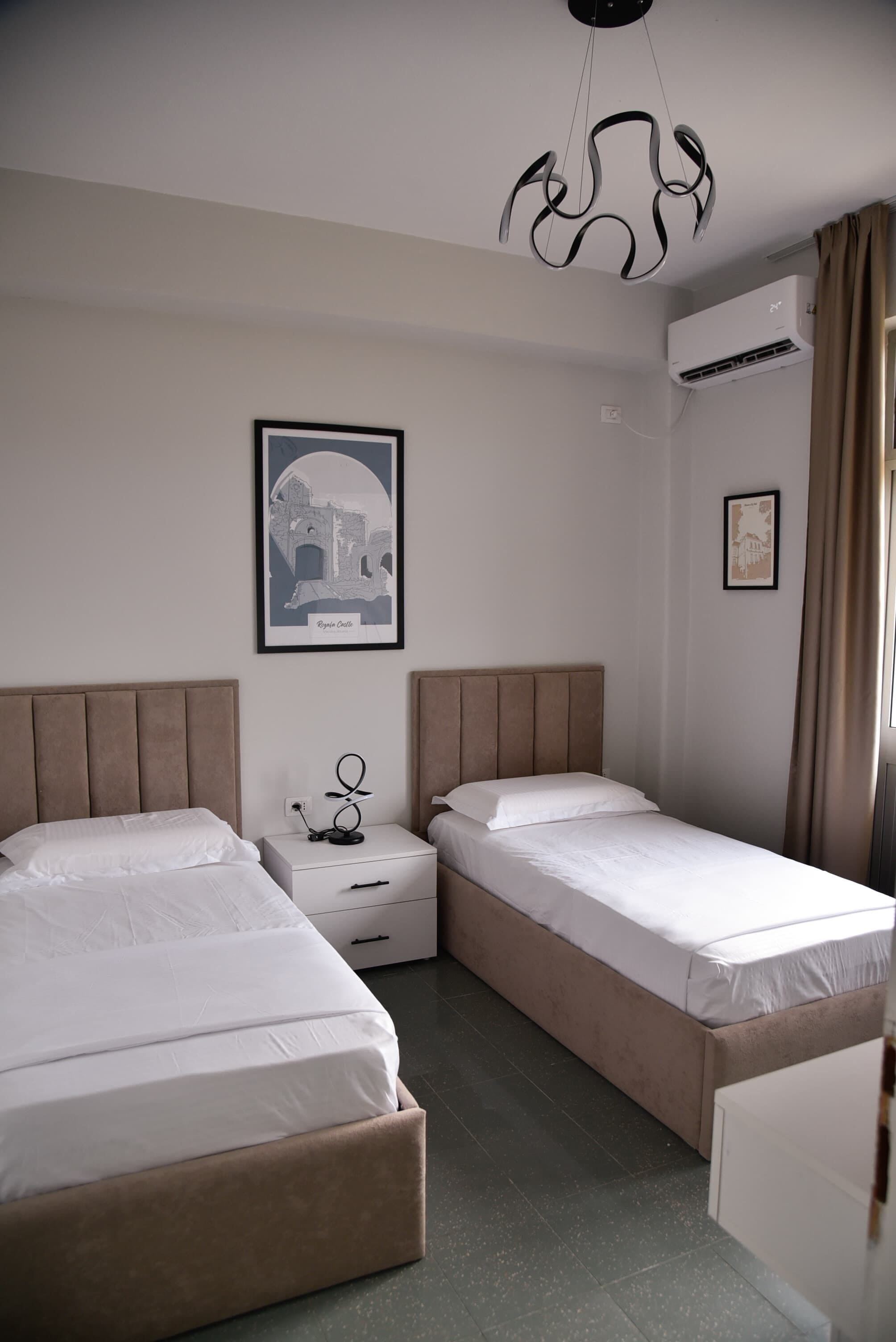 A hotel room with two beds, a nightstand, a framed picture on the wall, an air conditioner, and a window with curtains.