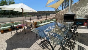 Een terras buiten met een glazen tafel, stoelen, een bank en een barbecue.