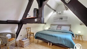 Un dormitorio con techo inclinado, una cama con cubrecama azul, una silla de madera, una mesita con lámpara y un estante en la pared con libros y un objeto decorativo.