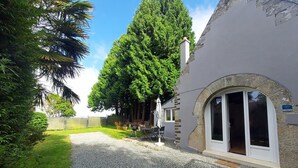 Un edificio con camino de grava, una zona de estar al aire libre cubierta y un gran árbol al fondo.