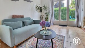Salón con sofá, una mesa de centro con flores, alfombra estampada y vista a un jardín por una puerta de vidrio.