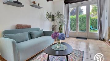 A living room with a sofa, a coffee table with flowers, a patterned rug, and a view through a glass door to a garden.