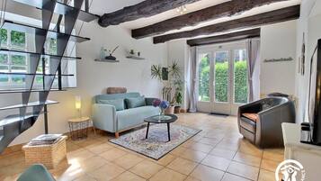 A modern living room with a sofa, a coffee table, and a fireplace. There is a staircase with glass railings and a view of greenery outside.