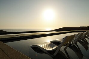 Zona de piscina con sillones de descanso y vista al océano al atardecer.