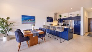 A modern kitchen with a central island, bar stools, and a dining table with chairs. - 2BR/2BA Oceanfront Lux Surf Condo in Los Cerritos w/ Pool, Gym, Hot Tub, Sauna  (Los Cerritos)