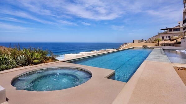 A pool area with a hot tub, a view of the ocean, and a hillside with buildings.