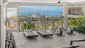 A gym with treadmills, free weights, and exercise machines, overlooking a beach and mountains. - 2BR/2BA Oceanfront Lux Surf Condo in Los Cerritos w/ Pool, Gym, Hot Tub, Sauna  (Los Cerritos)