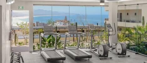 A gym with treadmills, free weights, and exercise machines, overlooking a beach and mountains.