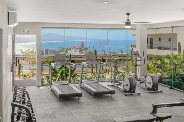 Un gimnasio con cintas de correr, pesas libres y máquinas de ejercicio, con vistas a una playa y montañas.