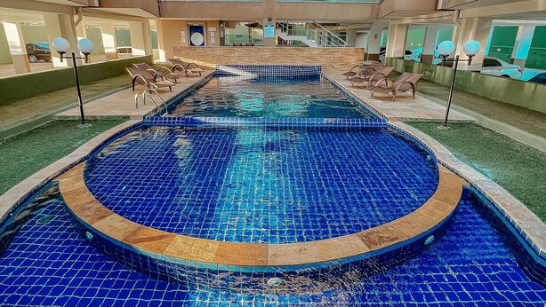 A large indoor swimming pool with a blue tiled floor, surrounded by lounge chairs and a multi-level building with balconies. - Apartamento Praia do Cumbuco (Caucaia)