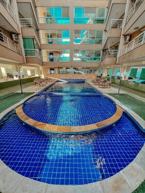 A large indoor swimming pool with a blue tiled floor, surrounded by lounge chairs and a multi-level building with balconies. - Apartamento Praia do Cumbuco (Caucaia)