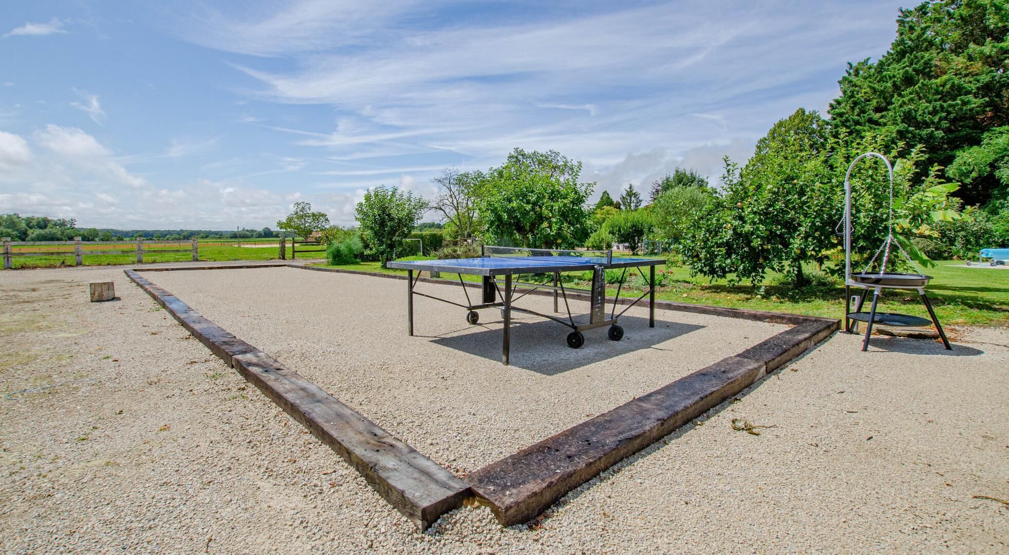 An outdoor ping pong table set up on a gravel surface with a surrounding wooden border, trees, and a swing set in the background.