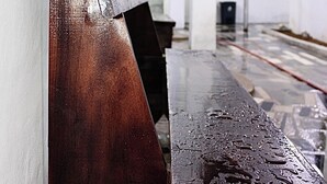 A wet wooden bench in a corridor with white walls and a tiled floor.