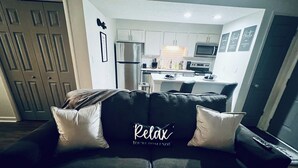 A modern living room with a black sofa and two white pillows, a kitchen in the background, and a framed picture on the wall. - Harmonious Character & Comfort (Huntsville)