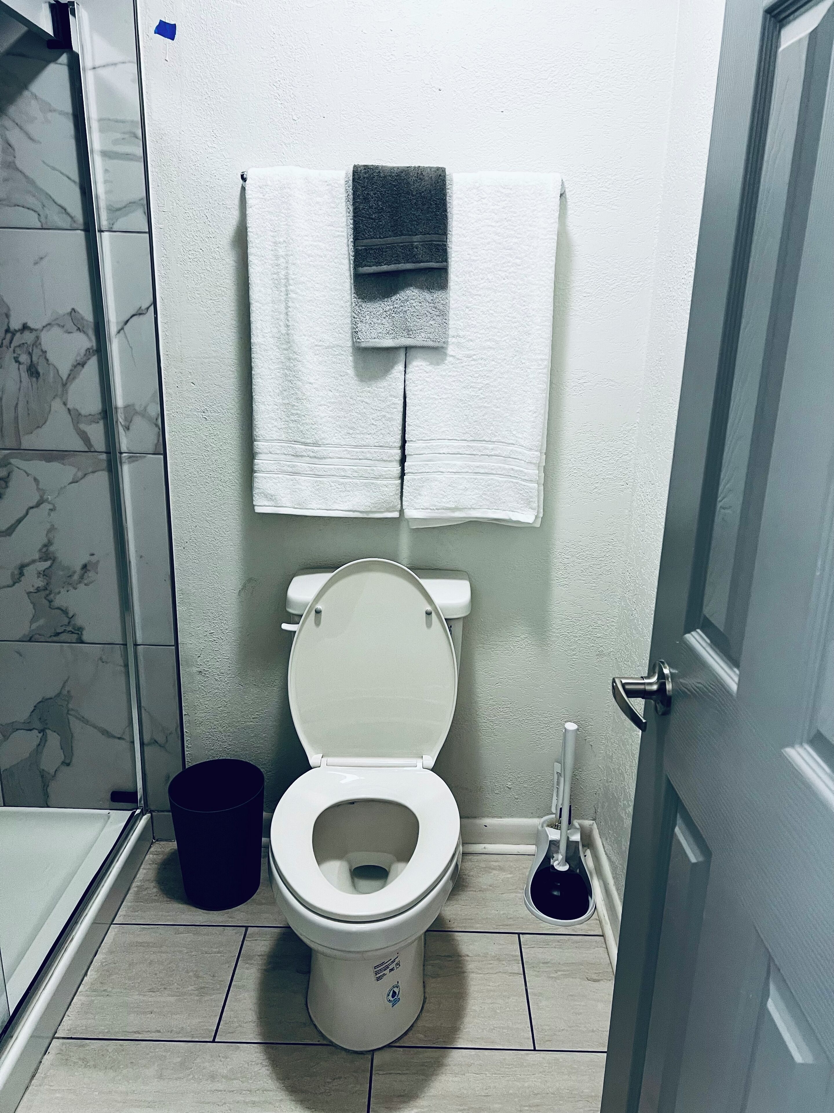 A bathroom with a toilet, a towel hanging on the wall, a plunger, and a bucket.