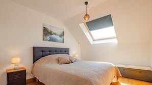A bedroom with a large bed, a skylight, a nightstand with a lamp, and a painting on the wall.