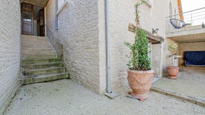 A stone building with a staircase leading to a door, a large terracotta pot with a plant, and a covered area with a chair.