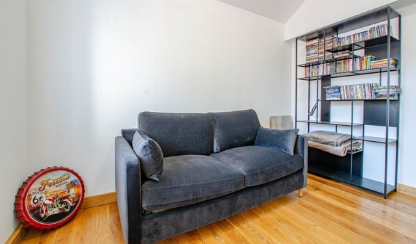 A grey sofa with cushions, a bookshelf filled with books and DVDs, and a red bottle cap with a motorcycle design on the floor.