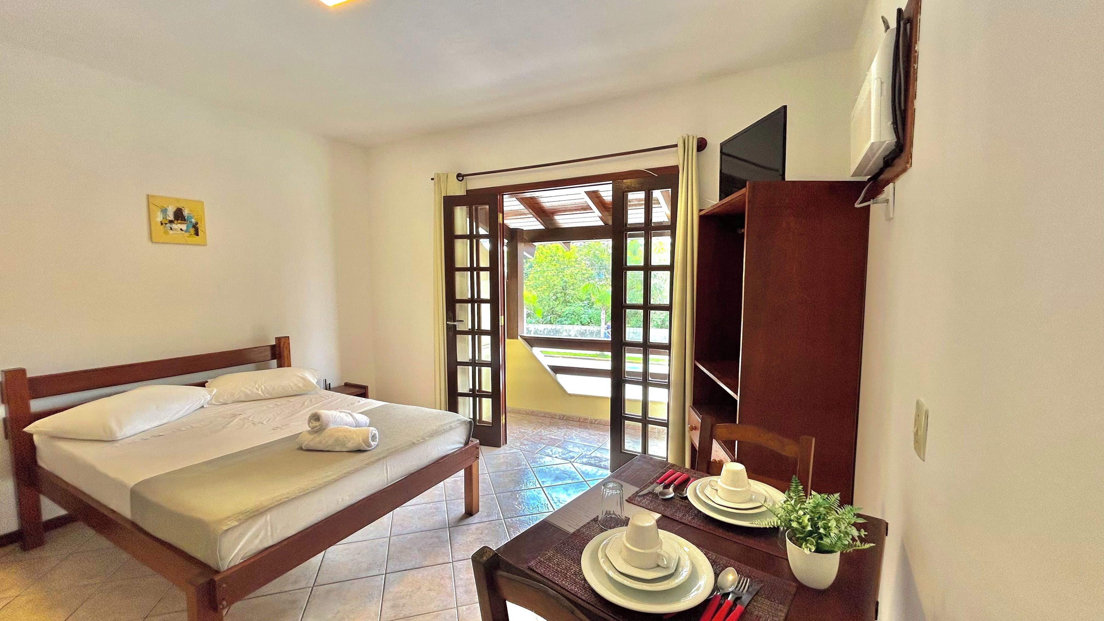Habitación de hotel con una cama, una mesa de comedor con platos, una planta y un televisor montado en la pared.