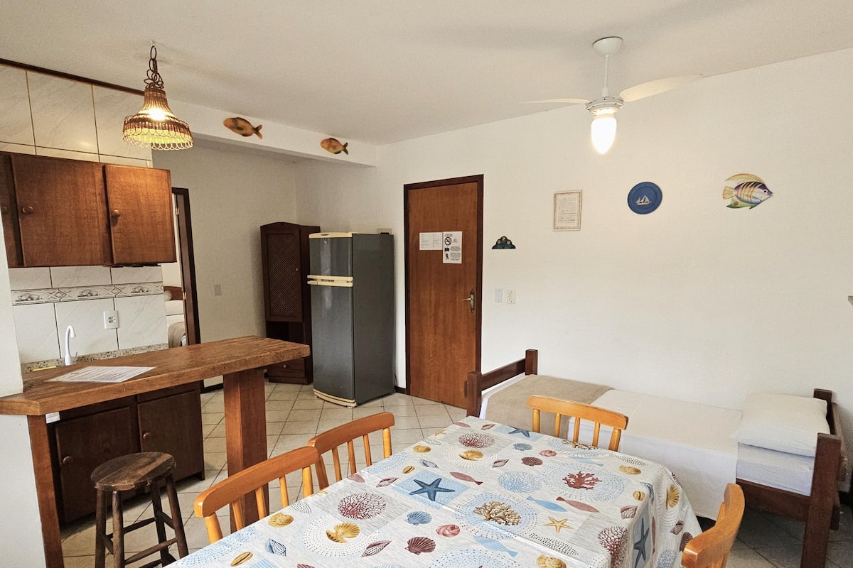 Egy praktikus, kisebb méretű konyha, melyben fa szekrények, egy étkezőasztal székekkel, egy hűtőszekrény és egy mennyezeti ventilátor található.