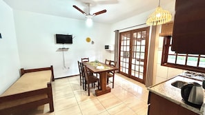Un comedor con mesa y sillas de madera, un ventilador de techo, un televisor montado en la pared y una cocina con lavamanos y estufa.
