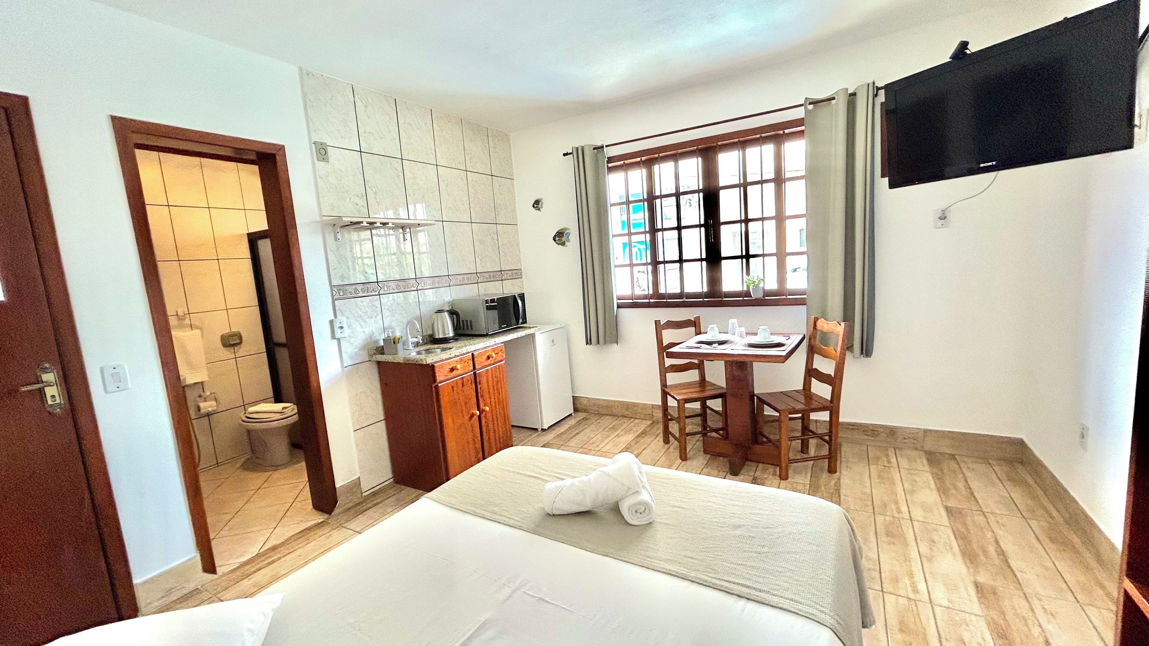 A hotel room with a bed, a small kitchenette, a dining table with chairs, a TV mounted on the wall, and a window with curtains.