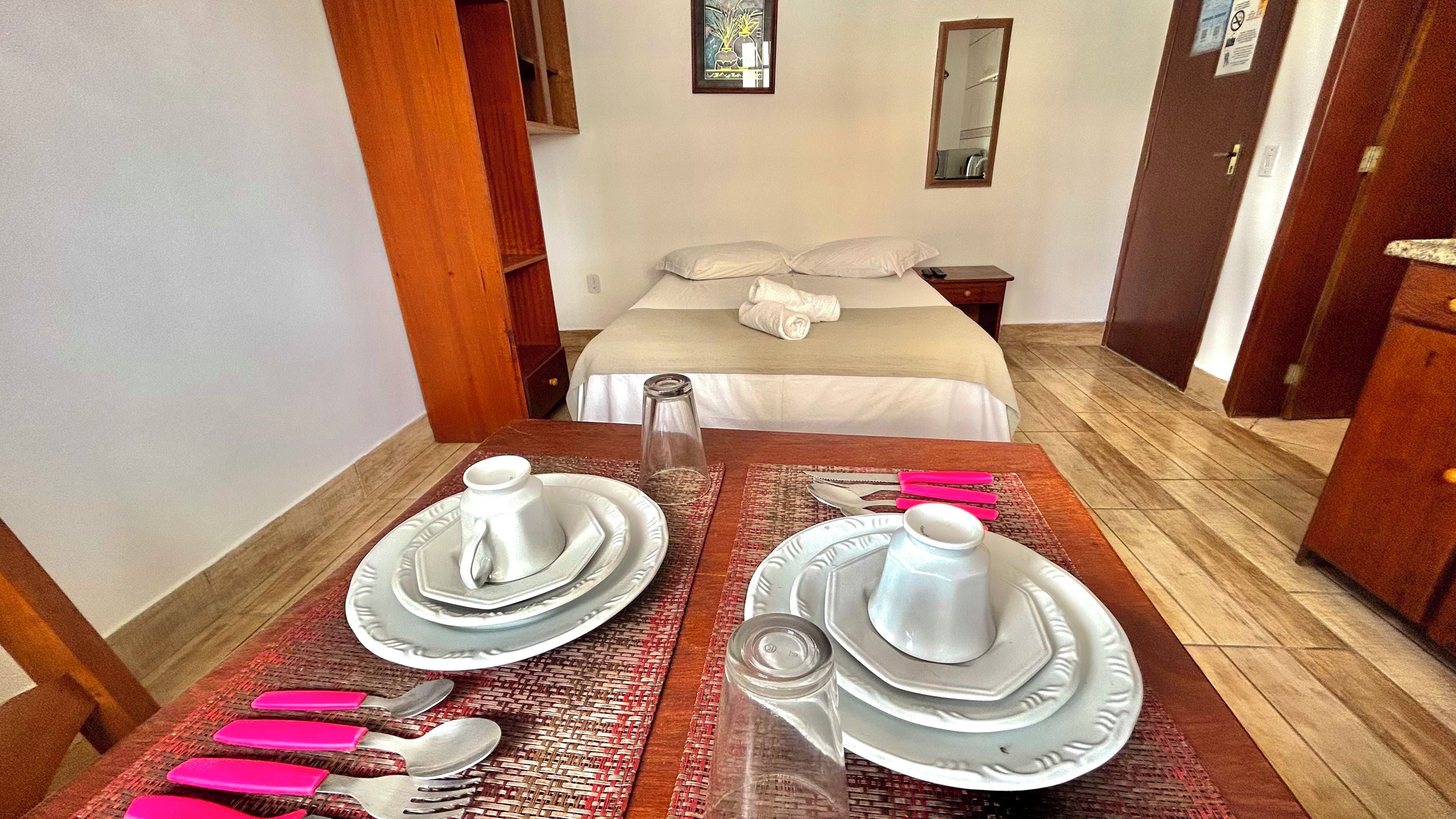 Quarto de hotel com cama bem arrumada, mesa de jantar posta com pratos, talheres e copos, e um pequeno objeto decorativo sobre a mesa.