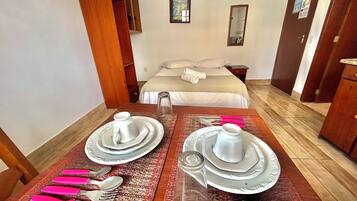Quarto de hotel com cama bem arrumada, mesa de jantar posta com pratos, talheres e copos, e um pequeno objeto decorativo sobre a mesa.