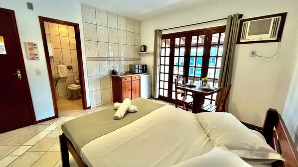 A hotel room with a bed, a desk, a view through a window, and a bathroom. - Pousada Jardim das Gales Bombinhas (Bombinhas)