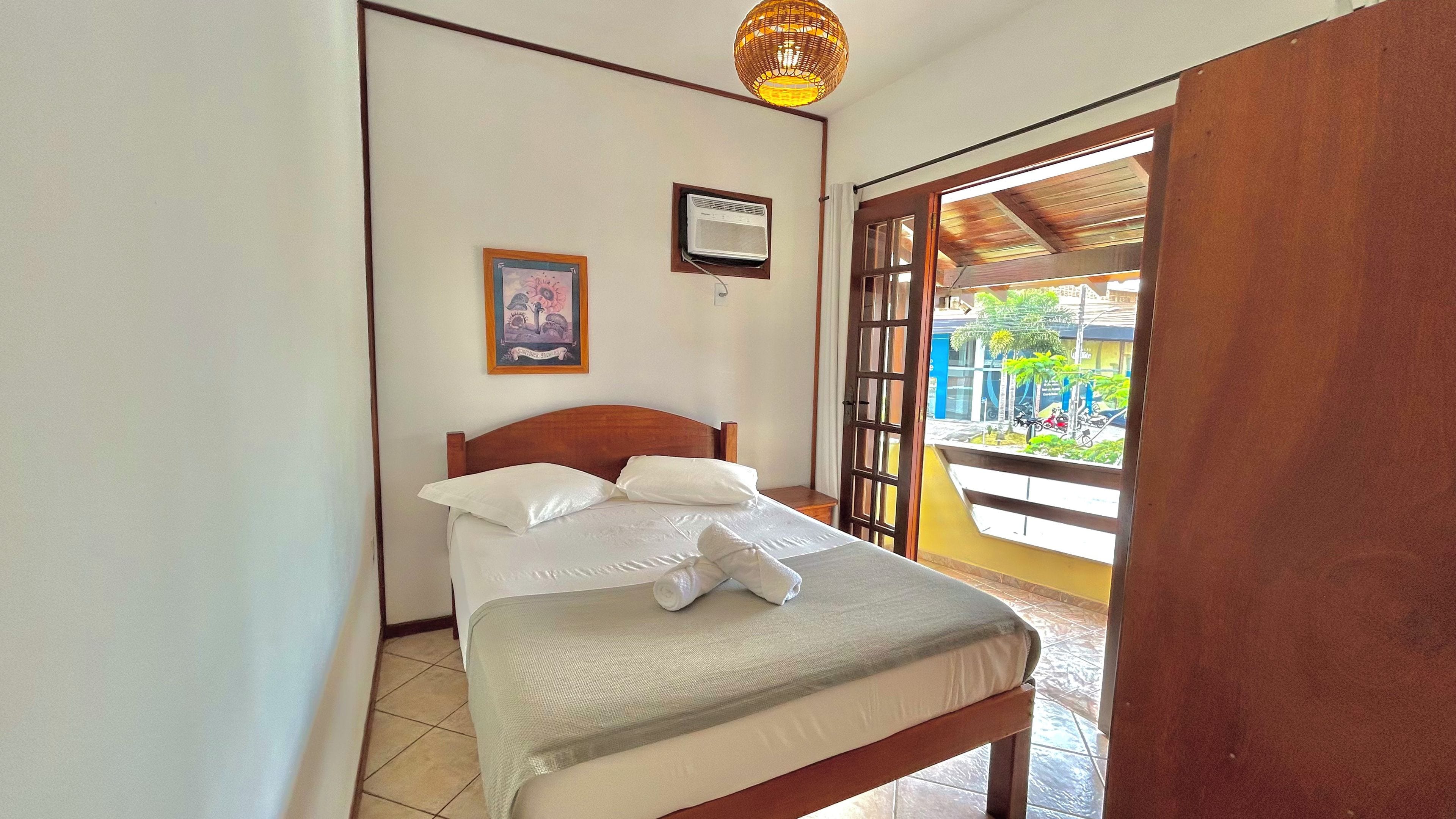 Un dormitorio con cama, aire acondicionado de pared, un cuadro enmarcado y vistas a través de una puerta abierta.