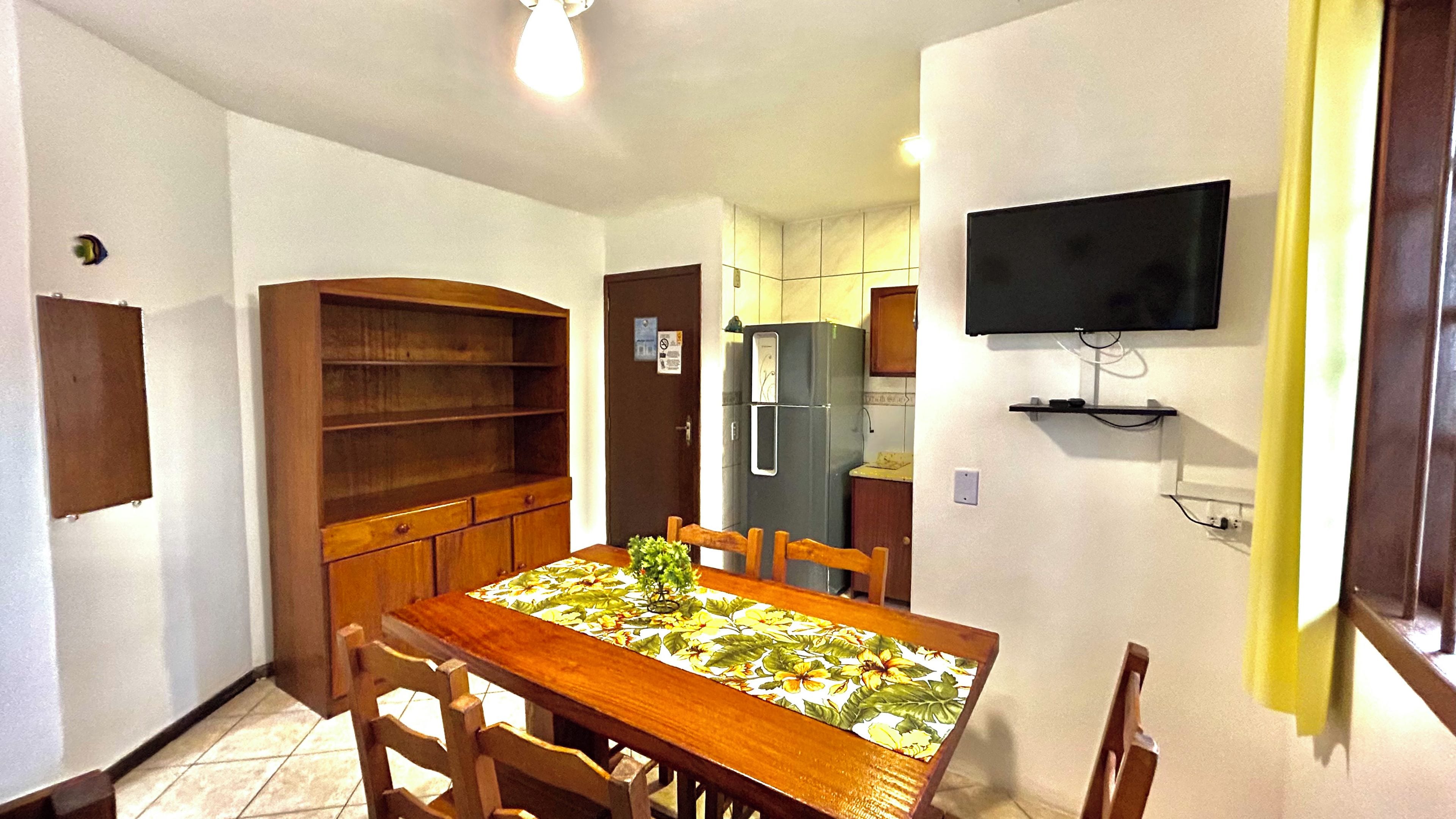 Una cocina compacta con una mesa de comedor, sillas, un armario de madera empotrado, un refrigerador y un televisor montado.