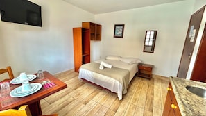 Habitació d'hotel amb llit, taula de nit, prestatge de fusta, taula de menjador amb plats i coberts, i televisió penjada a la paret.