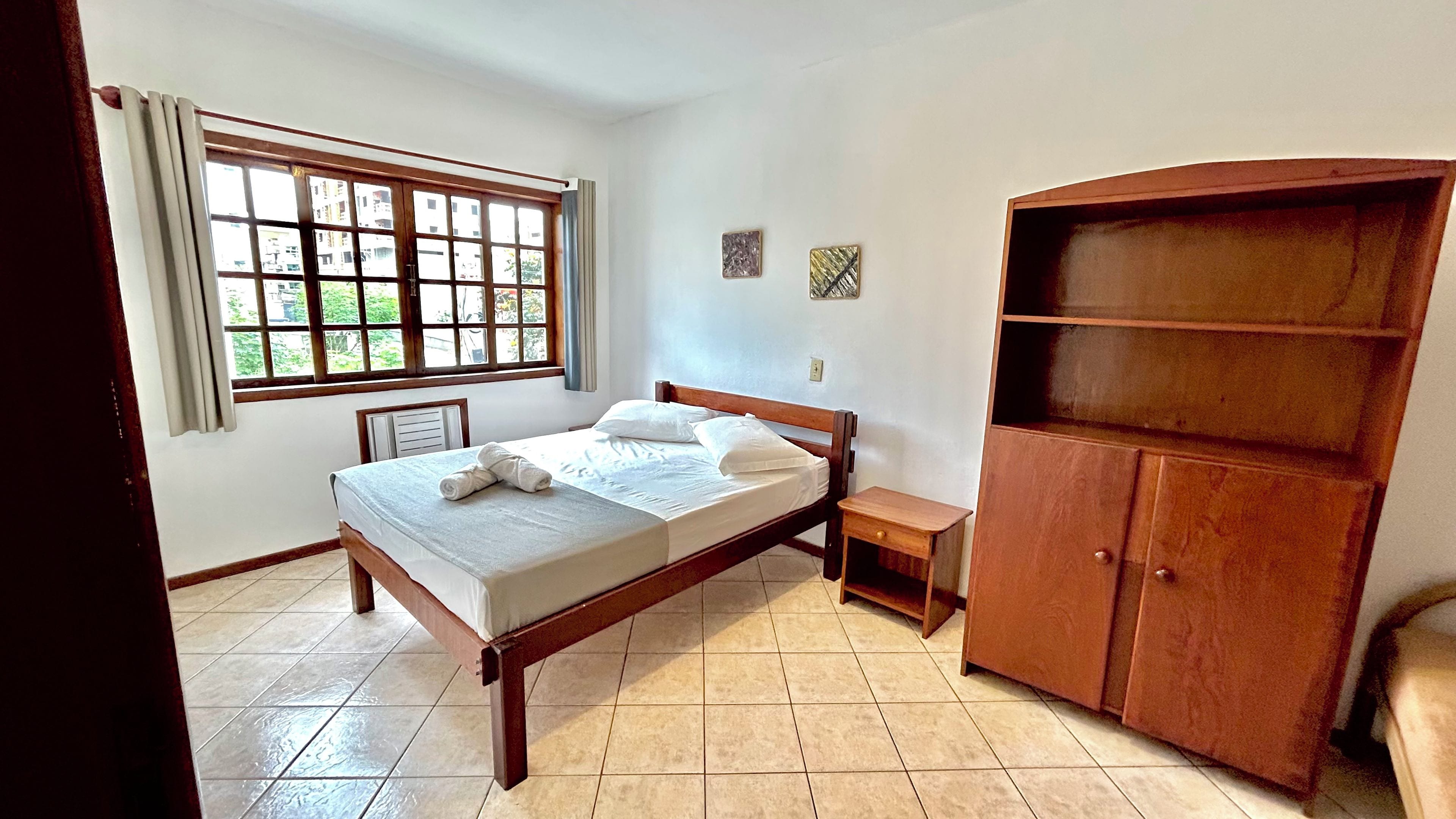 Un dormitorio con cama, un armario de madera, una mesita de noche y una ventana con vistas a edificios.