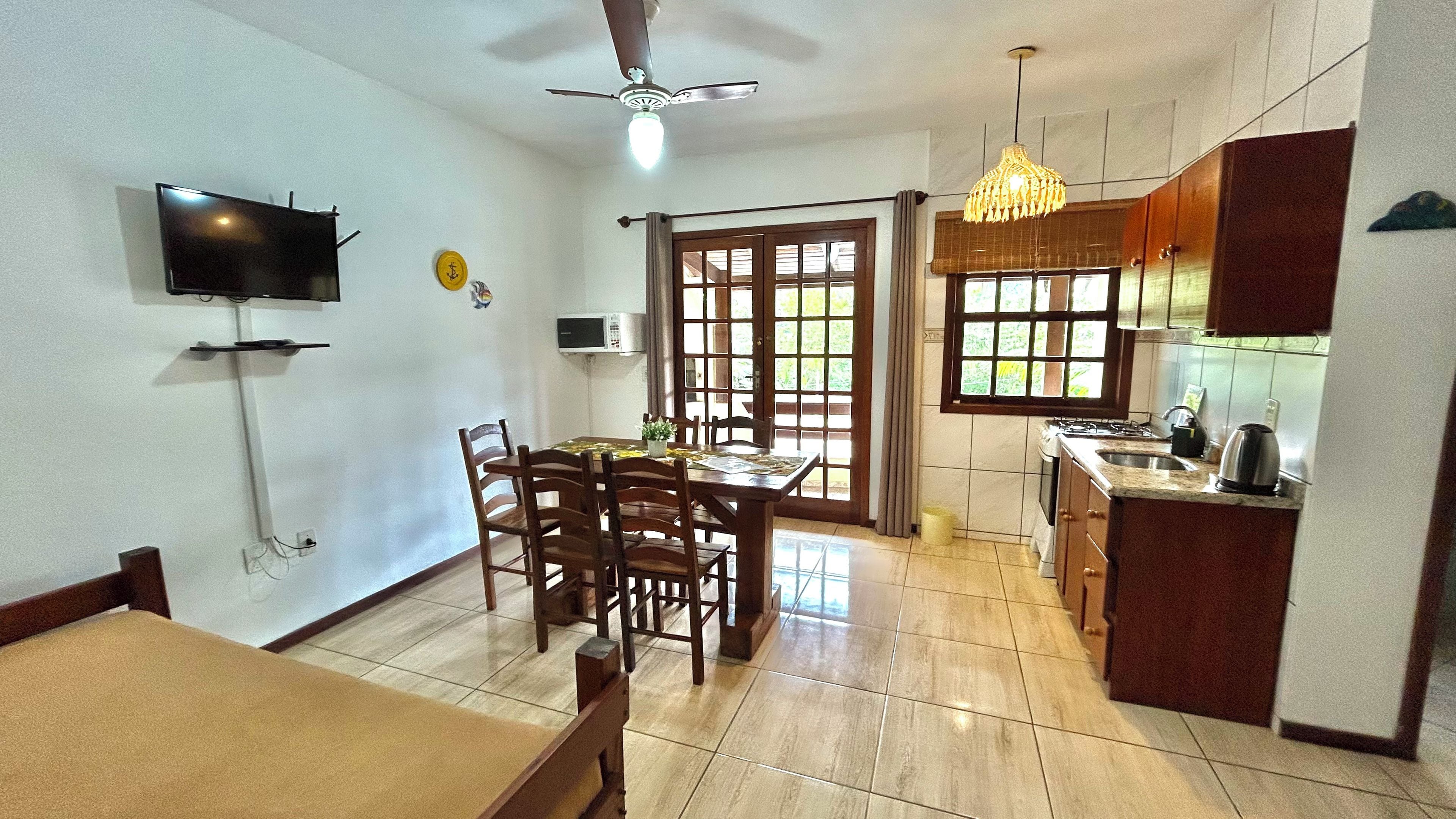 Un coin repas avec une table et des chaises, un téléviseur fixé au mur, un ventilateur de plafond et un espace cuisine équipé d’une cuisinière et d’un évier.