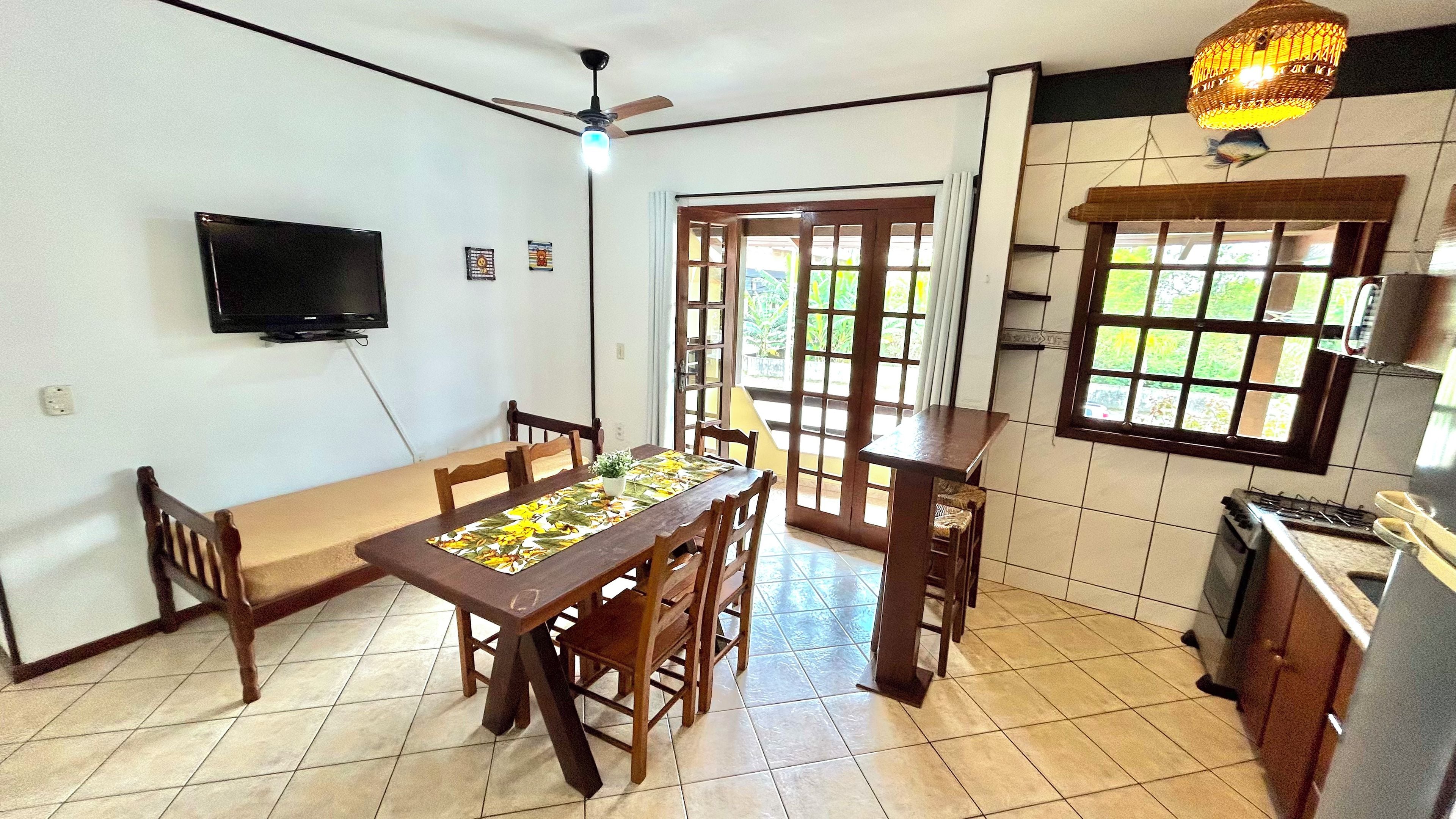 Un comedor con una mesa, sillas, ventilador de techo, televisor y ventana con vista a follaje.