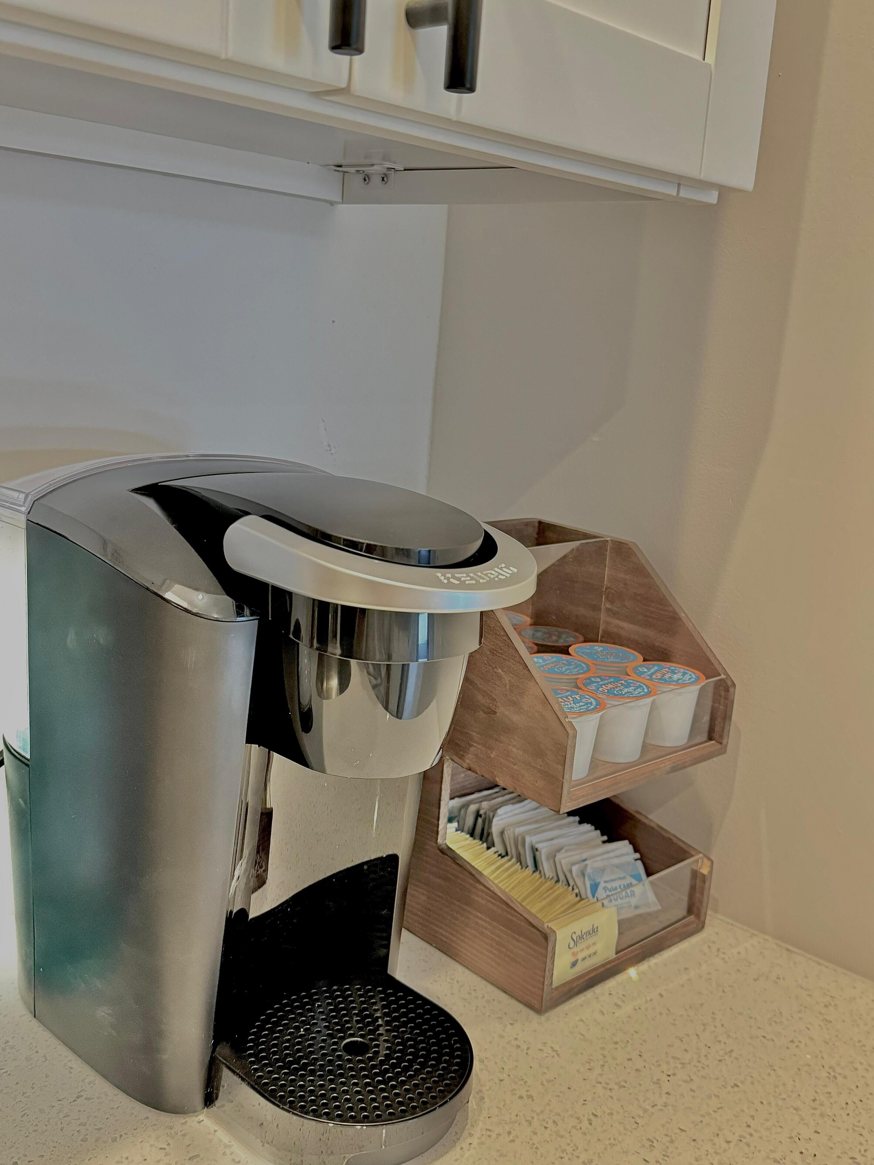 Una máquina de café con bandeja de goteo, un depósito para cápsulas de café y un recipiente para azúcar sobre una encimera.