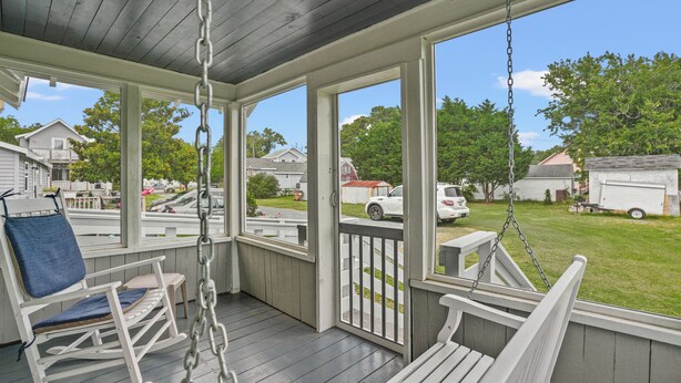 A porch with a swing chair and a rocking chair, a view of a residential street, and a grassy yard.