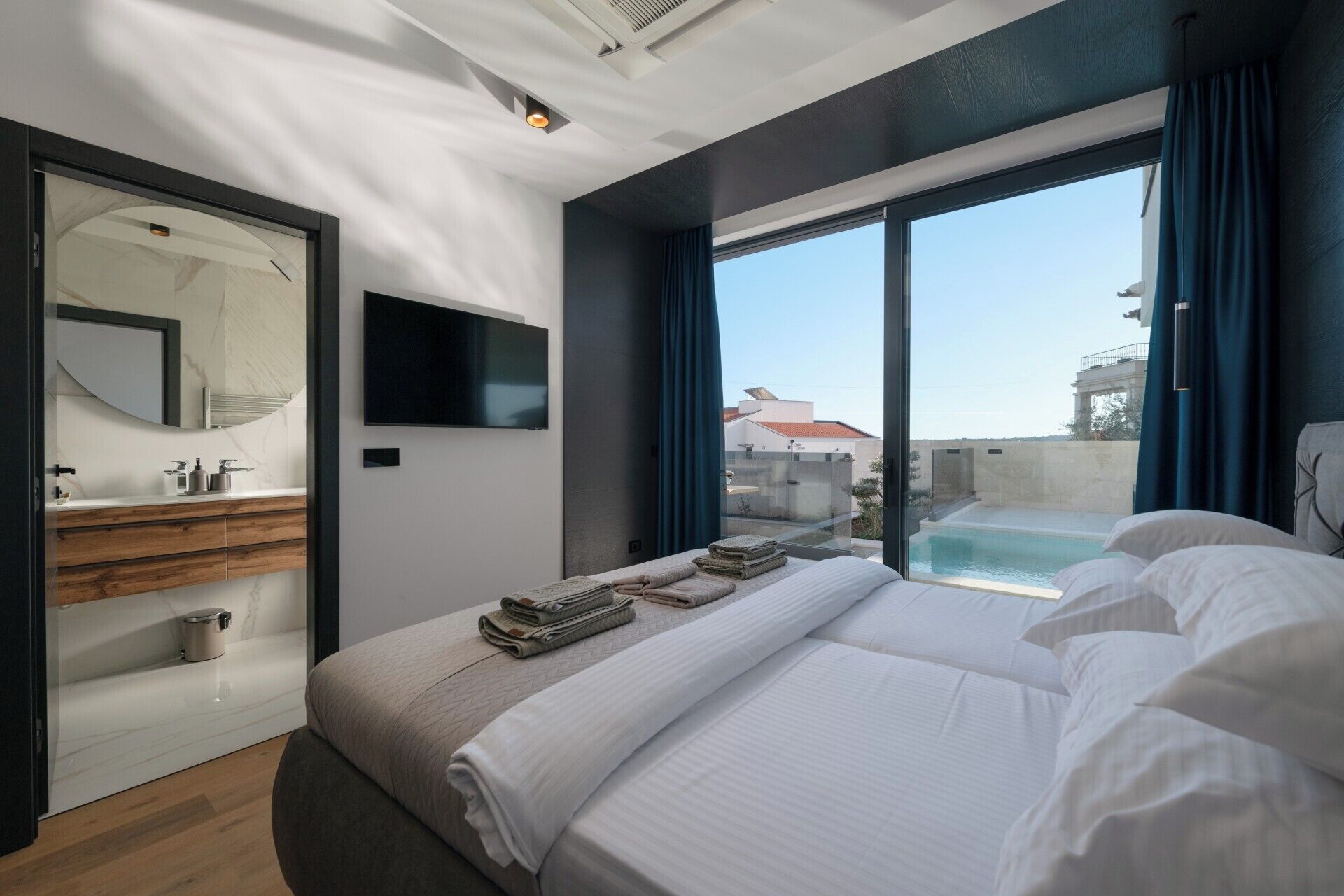 A modern hotel room with a large bed, a TV mounted on the wall, a bathroom with a mirror and sink, and a view of a pool and buildings outside.