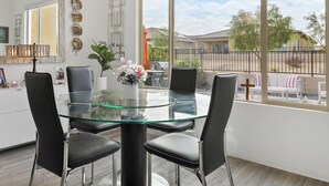 A modern dining area with a glass table and black chairs, a pendant light, and a view of a residential neighborhood through large windows. - 2-bedroom house in superb Rancho Mirage Dell Webb 55+ Community. (Rancho Mirage)