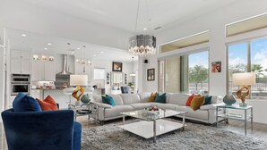 A modern living room with a white sectional sofa, a blue armchair, and a glass coffee table. There is a kitchen in the background with white cabinets and stainless steel appliances. - 2-bedroom house in superb Rancho Mirage Dell Webb 55+ Community. (Rancho Mirage)