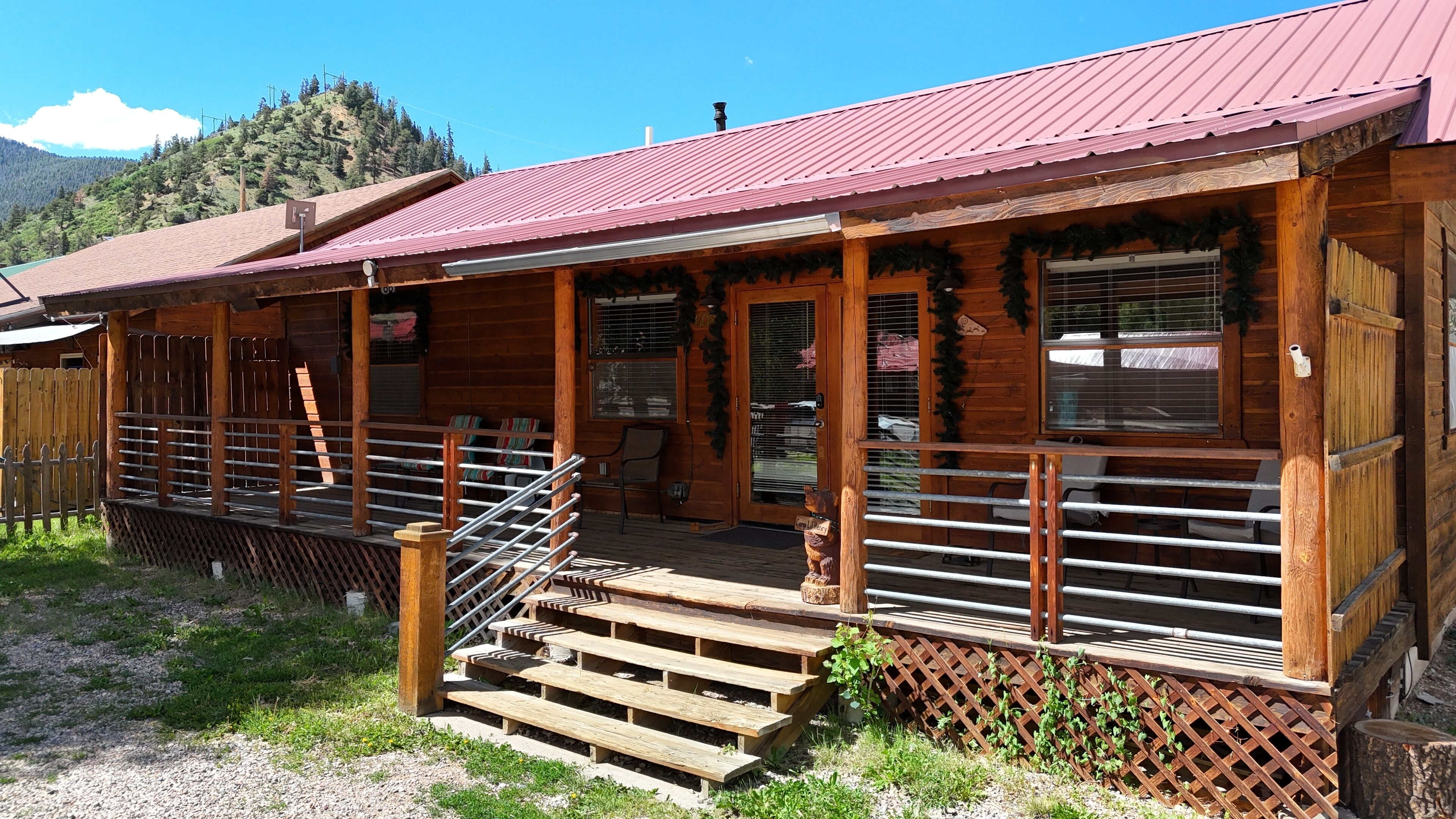 A wooden cabin with a red roof, a porch with steps, and a fenced area.