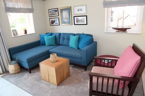 Una sala de estar con un sofá seccional azul, una mesa de centro de madera, una silla de madera y una mesita con un cojín rojo.