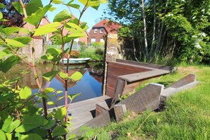 A wooden deck with a pond, surrounded by greenery and a brick building in the background. - Framheim - 100qm Luxus-ferienhaus für 7 Personen Direkt am Wasser bei Greetsiel! (Krummhörn)