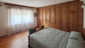 A bedroom with a wooden wardrobe, a bed with patterned bedding, a chair, and a window with sheer curtains. - Apartment 'Appartamento Nide A Vodo' with Mountain View, Private Terrace and Balcony (Vodo di Cadore)