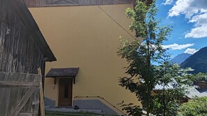 A building with a sloped roof, a small attached structure with a door, and a grassy yard with a bench. - Apartment 'Appartamento Nide A Vodo' with Mountain View, Private Terrace and Balcony (Vodo di Cadore)